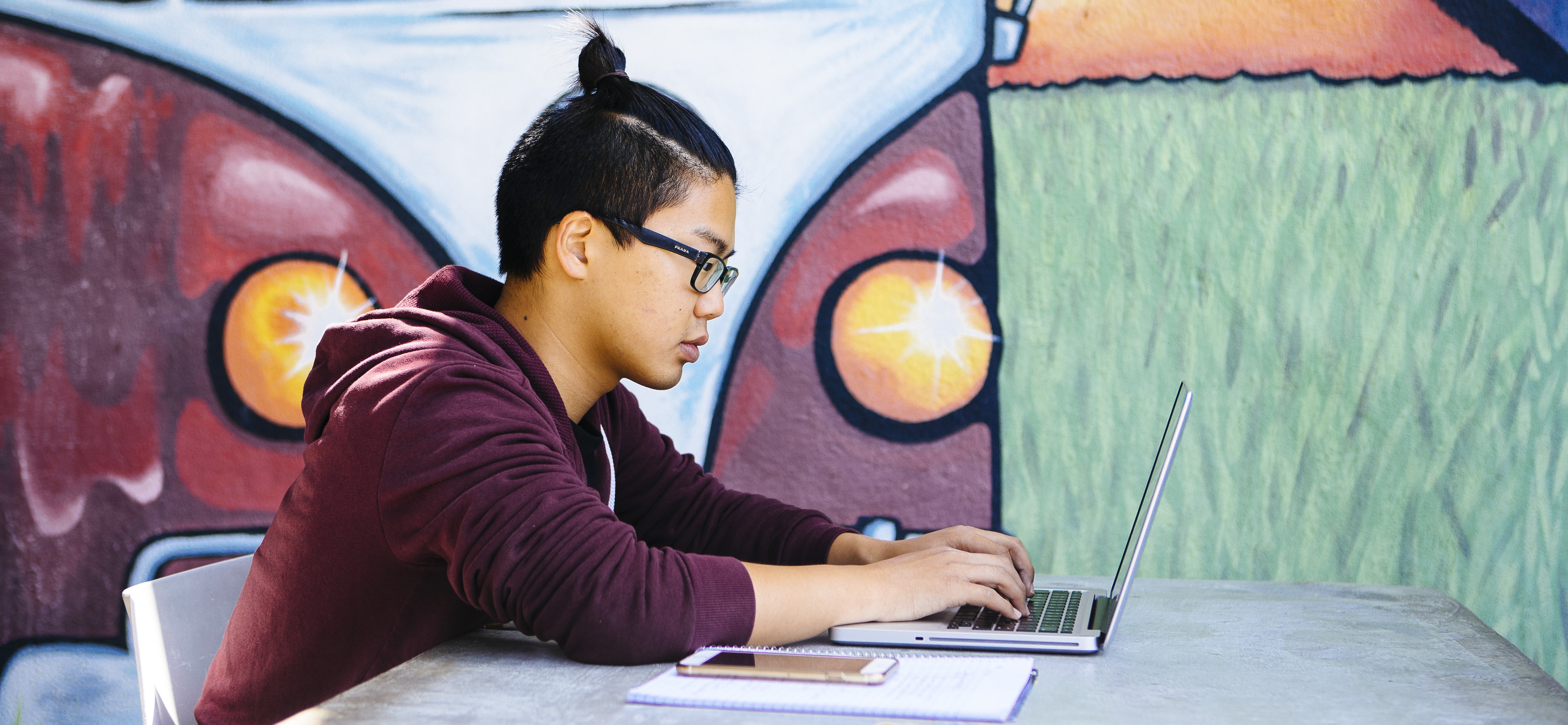 An asian student with glasses types on a laptop in front of a mural.